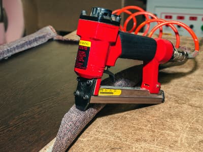 Ein Druckluft-Tacker fixiert Stoff auf einer Holzplatte.
