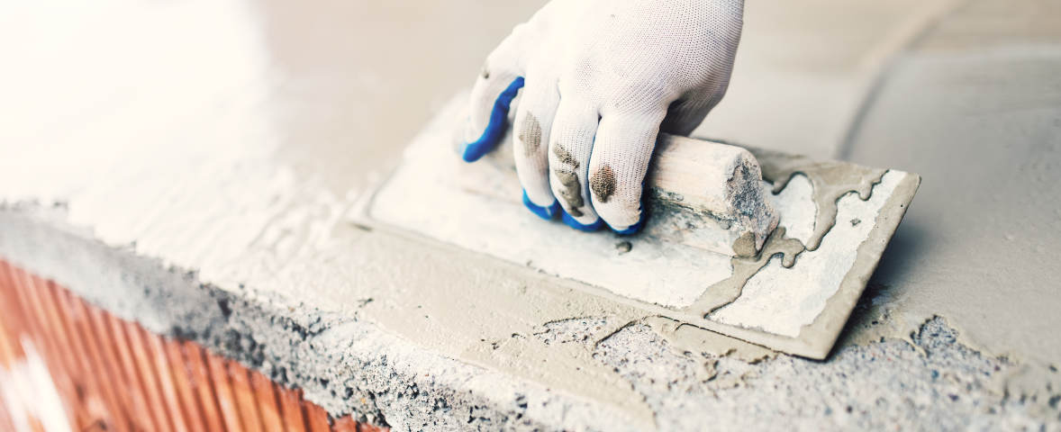 Eine Hand mit Arbeitshandschuhen glättet mit einer Kelle frischen Putz oder Mörtel auf einer gemauerten Fläche. Der Baustoff wird gleichmäßig verteilt.