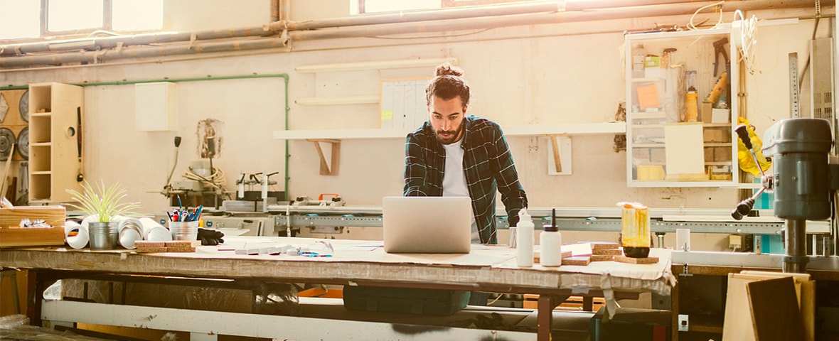 Ein Mann steht in einer gut ausgestatteten Werkstatt an einem großen Arbeitstisch und arbeitet an einem Laptop. Auf dem Tisch liegen Werkzeuge, Pläne und verschiedene Materialien.