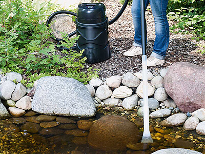 Eine Person reinigt einen Gartenteich mit einem Teichsauger. Der schwarze Sauger steht am Ufer auf einer mit Rindenmulch bedeckten Fläche, der Saugschlauch wird im flachen Wasser zwischen Steinen verwendet. Um den Teich herum liegen große Natursteine und wachsen verschiedene Pflanzen