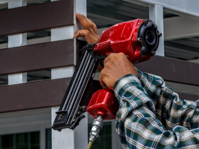 Eine Person befestigt Holzbalken mit einem Druckluftnagler an einem Metallgerüst.