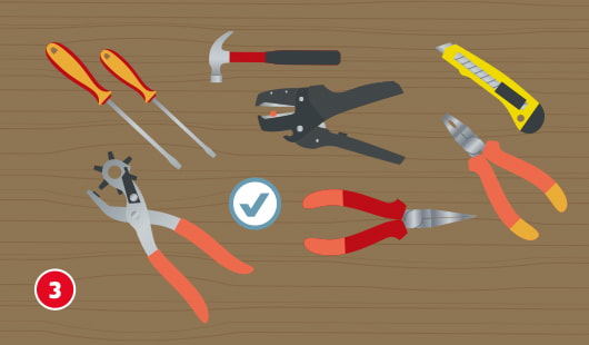 Eine schematische Darstellung zeigt eine Auswahl an Werkzeugen auf einer Holzfläche, die für den Bau eines Lampenschirms benötigt werden. Abgebildet sind zwei Schraubendreher, ein Hammer, eine Abisolierzange, ein Cuttermesser, eine Kombizange, eine Lochzange und eine Spitzzange. Ein Häkchensymbol kennzeichnet die Zusammenstellung als geeignet. Unten links ist ein roter Kreis mit einer weißen 3