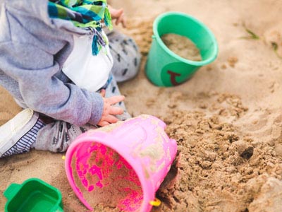 Ein kleines Kind spielt im Sandkasten mit buntem Sandspielzeug. Im Bildvordergrund liegt ein umgekippter pinker Eimer mit Sandresten, daneben stehen ein grüner Eimer und eine grüne Sandform