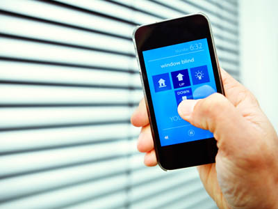 Eine Person steuert per Smartphone-App einen Rollladen mit den Funktionen Hoch und Runter vor einem Fenster mit Lamellen