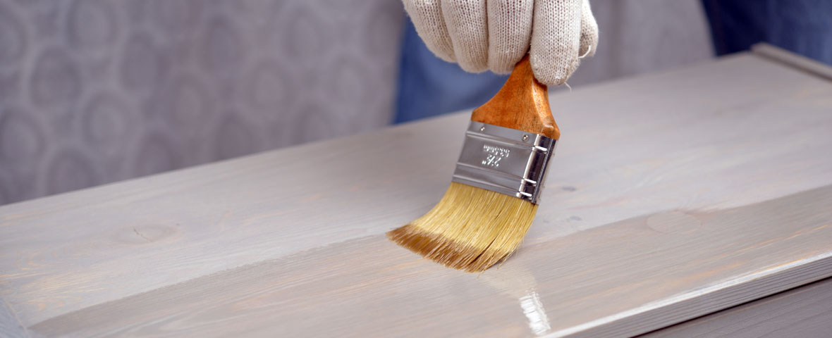 Detailaufnahme von Oberfläche einer weißen Kommode, auf der mit einem Pinsel Lack aufgetragen wird