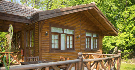 Ein großes Gartenhaus aus Holz mit mehreren Fenstern und einer überdachten Terrasse steht inmitten grüner Bäume. Das Haus wirkt wohnlich und ist mit einem Ziegeldach gedeckt