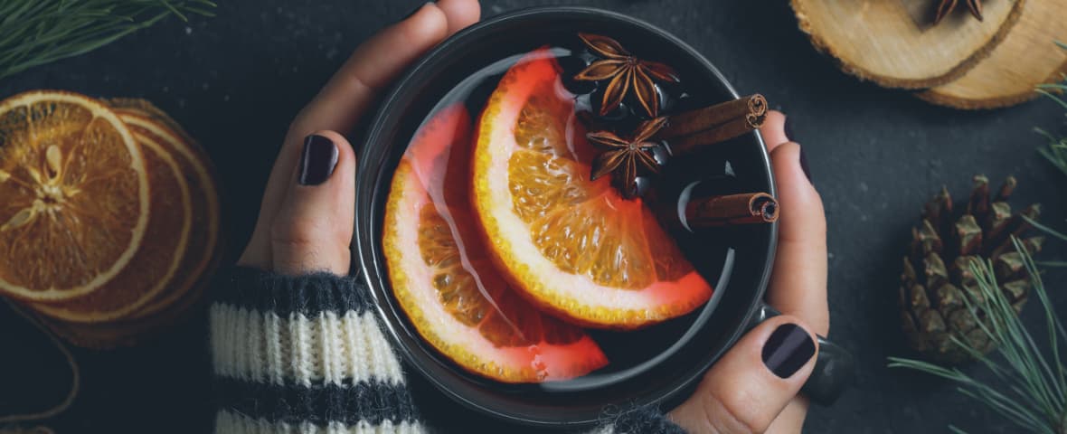 Zwei Hände mit dunkel lackierten Fingernägeln und gestreiften Wollärmeln halten eine Tasse Glühwein mit Orangenscheiben, Zimtstangen und Sternanis. Daneben liegen getrocknete Orangenscheiben, Holzscheiben, Tannenzweige und ein Tannenzapfen auf dunklem Untergrund