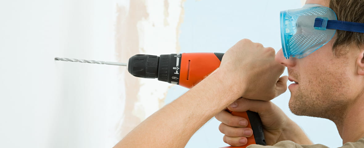 Ein Mann mit Schutzbrille bohrt mit einer orangefarbenen Bohrmaschine in eine weiße Wand. Dabei hält er das Gerät konzentriert mit beiden Händen.