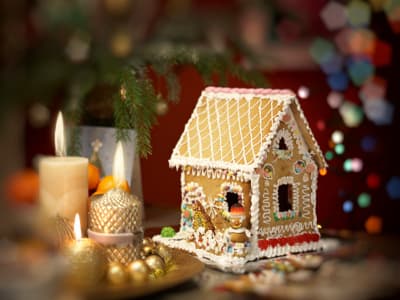 Ein kunstvoll verziertes Knusperhäuschen aus Lebkuchen steht auf einem weihnachtlich dekorierten Tisch. Das Haus ist mit weißem Zuckerguss, bunten Süßigkeiten und Schokoladentäfelchen geschmückt. Daneben brennen Kerzen in goldenen Haltern, Kugeln und Tannengrün runden die Dekoration ab. Im Hintergrund sind bunte Lichter unscharf zu erkennen