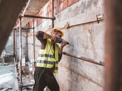 Ein Bauarbeiter mit Schutzhelm und Warnweste bohrt mit einer Schlagbohrmaschine in eine Betonwand. Er arbeitet auf einer Baustelle zwischen Gerüsten.