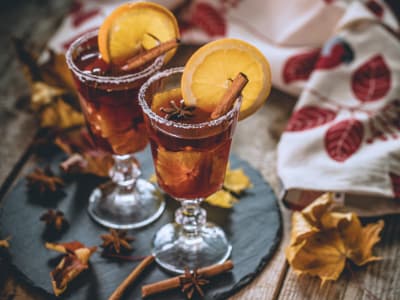 Zwei Gläser mit Glühbier stehen auf einer dunklen Schieferplatte. Das Getränk ist mit Orangenscheiben, Zimtstangen und Sternanis dekoriert, der Glasrand ist mit Zucker verziert. Rundherum liegen herbstliche Blätter, Gewürze und ein besticktes Tuch mit roten Blättermotiven auf einem Holztisch