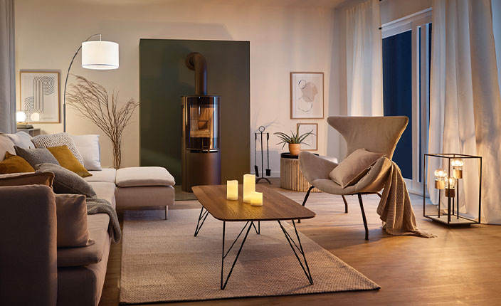 Ein gemütlich eingerichtetes Wohnzimmer ist mit mehreren Lichtquellen in warmem Licht gestaltet. Auf dem Couchtisch stehen flammenlose Kerzen, in der Ecke leuchtet ein großer Kerzenständer mit Echtwachskerzen. Eine Stehlampe mit weißem Schirm, indirekte Beleuchtung und ein Kaminofen sorgen zusätzlich für stimmungsvolles Licht. Die Einrichtung ist modern und natürlich gehalten