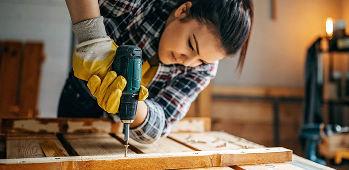 Eine Frau schraubt mit einer Bohrmaschine ein Holzbalken an Holzbrettern fest