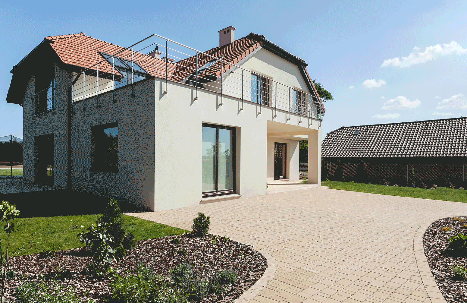 Eine große Terrasse hinter einem modernen Einfamilienhaus mit roten Dachziegeln. In einer Animation erscheinen nacheinander verschiedene Einrichtungsgegenstände wie Sitzmöbel, Tisch, Hängesessel, Sonnenschirm, Pflanztöpfe und ein Hochbeet als schwarz-weiße Zeichnungen. Oben links steht in weißer Schrift auf grünem Hintergrund: „Große Terrasse gemütlich gestalten“.