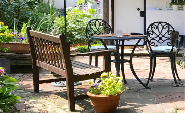 Eine Braune Bank die an einem kleinen schwarzen Gartentisch steht in einem Grünen Garten