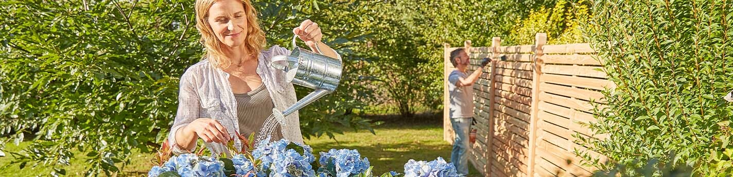 Eine Frau bewässert blaue Blumen und ein Mann streicht im Hintergrund einen Zaun
