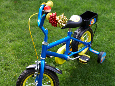 Ein kleines blaues Kinderfahrrad mit Stützrädern steht auf einer Rasenfläche. Am Lenker sind eine gelbe Klingel und zwei Geschenkschleifen befestigt, auf dem Gepäckträger ist eine schwarze Transportbox montiert