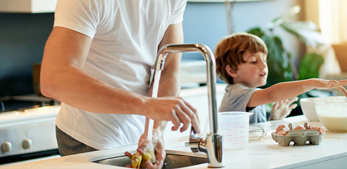 Ein Mann wäscht Gemüse unter einer modernen Küchenarmatur, während ein Kind beim Backen hilft.