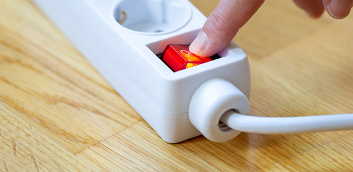 Hand schaltet den roten Kippschalter einer Mehrfachsteckdose auf einem Holzboden aus.