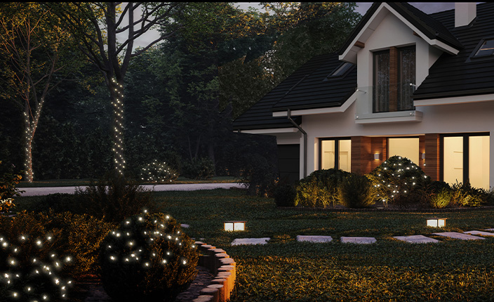 Ein Garten vor einem Einfamilienhaus ist mit mehreren Lichterketten dekoriert. Leuchtpunkte befinden sich auf rund geschnittenen Büschen, entlang eines geschwungenen Gartenwegs und spiralförmig um die Stämme einzelner Bäume gewickelt. Das Haus ist von innen beleuchtet