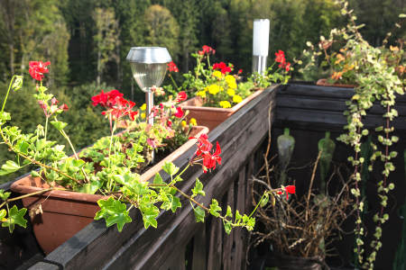 Eine bepflanzte Holzterrasse mit Balkonkästen voller roter und gelber Blumen. Zwischen den Pflanzgefäßen steht eine kleine Solarlampe zur dezenten Beleuchtung. Im Hintergrund ist ein bewaldeter Hang zu sehen