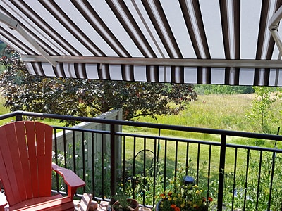 Blick von einem Balkon mit schwarzem Metallgeländer auf eine grüne Wiesenlandschaft. Über dem Balkon ist eine gestreifte Markise in Weiß und Dunkelbraun ausgefahren und spendet Schatten. Links steht ein roter Gartenstuhl, am Geländer hängen Blumenkästen mit bunten Pflanzen.