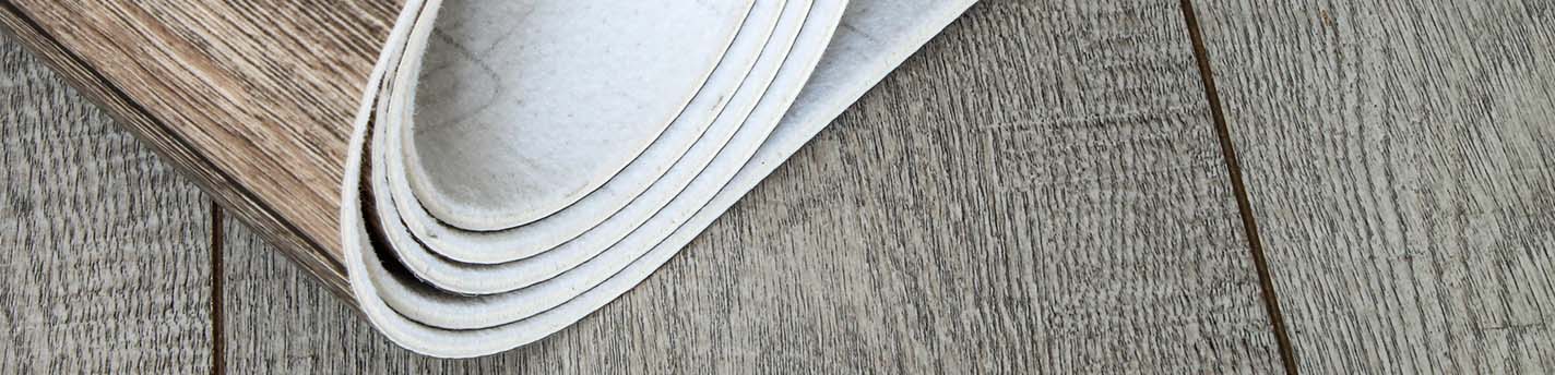 Aufgerollter Vinylboden in Holzoptik auf grauem Holzboden.