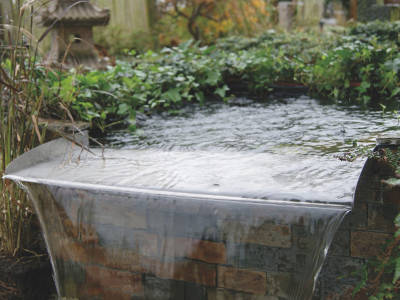 Ein moderner, überlaufender Gartenteich mit gerader Wasserkante aus Metall, über die das Wasser in einen darunterliegenden Bereich fließt. Der Teich ist von Kletterpflanzen und Natursteinen umgeben.