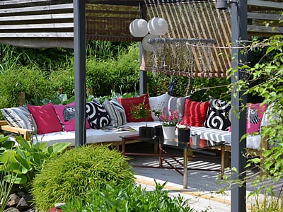 Eine Pergola mit Lamellendach in einem Garten, unter der Pergola stehen eine Sitzecke mit Polstern und Kissen und ein Tisch