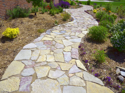 Ein geschwungener Gartenweg aus unregelmäßigen Natursteinplatten führt durch ein neu angelegtes Beet mit Mulch und blühenden Stauden