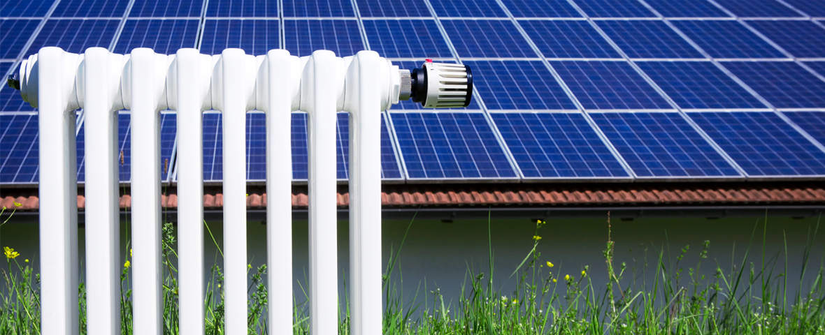 Ein weißer Heizkörper steht im Vordergrund, im Hintergrund ist ein mit Solarpaneelen bedecktes Hausdach zu sehen. Das Bild symbolisiert die Verbindung von Heizung und Solarenergie.
