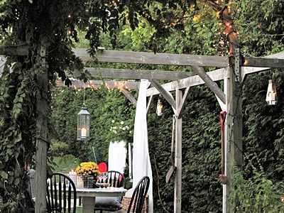 Romantisch dekorierte Pergola im Garten mit Holztisch und schwarzen Stühlen. Lichterketten, Laternen und Pflanzenranken schmücken die Holzkonstruktion, im Hintergrund dichte Heckenbepflanzung.