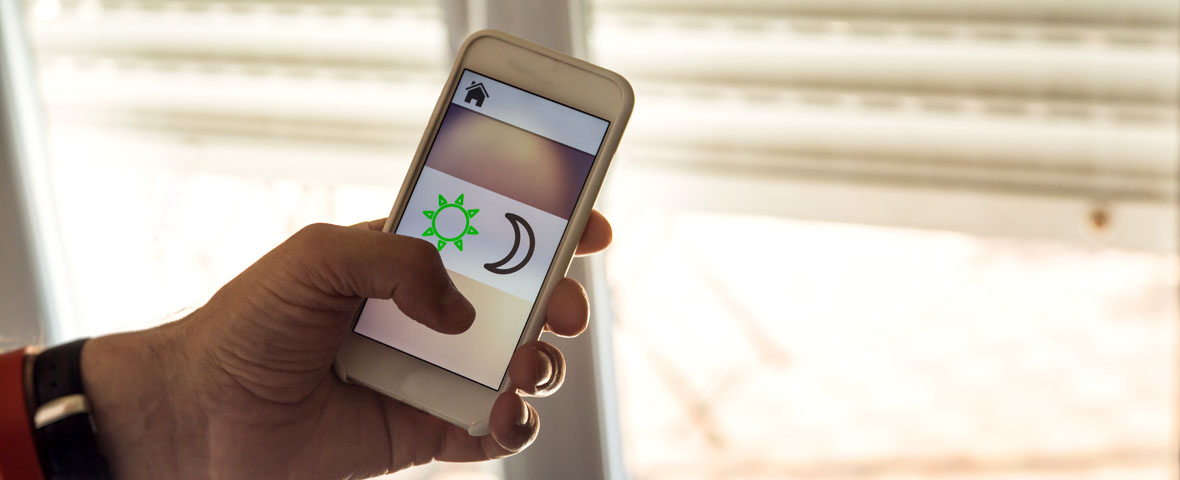 Eine Hand bedient per Smartphone-App die Rollladensteuerung mit Tag-und-Nacht-Funktion vor einem Fenster