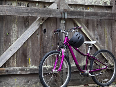 Ein pinkfarbenes Kinderfahrrad mit einem schwarzen Fahrradhelm am Lenker steht vor einem alten hölzernen Scheunentor. Das Fahrrad hat eine weiße Satteldecke und schwarze Reifen