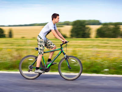 Ein Jugendlicher fährt auf einem grünen Fahrrad eine Landstraße entlang. Er trägt ein graues T-Shirt und gemusterte Shorts. Im Hintergrund ist eine weite Landschaft mit Feldern und Bäumen zu sehen