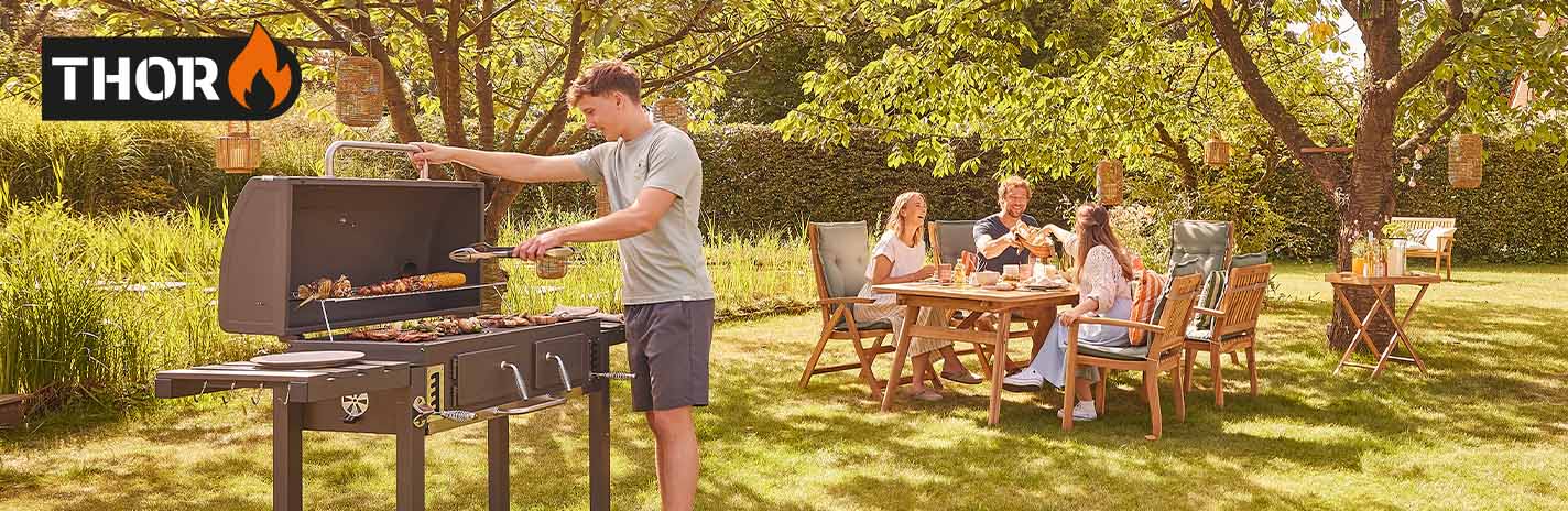 Ein junger Mann grillt etwas für seine Familie, die im Hintergrund auf Gartenmöbeln sitzen