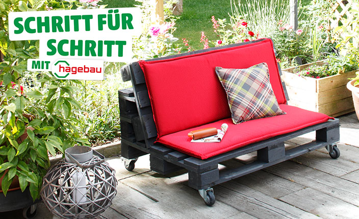 Selbstgebaute Gartenbank aus schwarzen Paletten mit roten Polstern und einem karierten Kissen, auf einer Terrasse mit Pflanzen im Hintergrund. Text: "Schritt für Schritt mit hagebau".