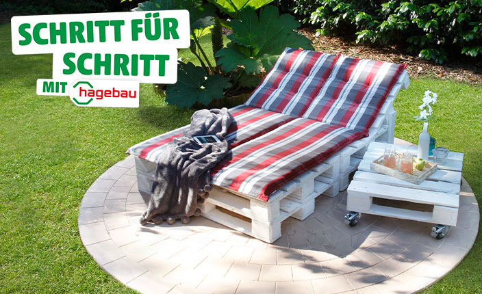 Gartenliege aus weißen Europaletten mit rot-weiß gestreiften Polstern und einer grauen Decke, daneben ein kleiner Paletten-Beistelltisch mit Getränken und Blumen, auf einer runden Terrasse im Grünen. Text: "Schritt für Schritt mit hagebau".