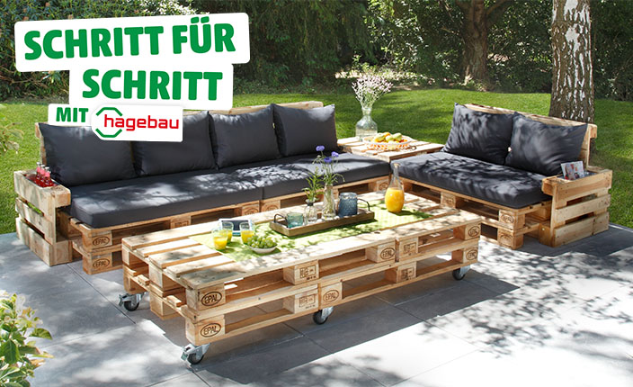 Gartenlounge aus Europaletten mit grauen Polstern und Kissen, bestehend aus Sofa, Sessel und Couchtisch auf Rollen, auf einer Terrasse im Grünen. Text: "Schritt für Schritt mit hagebau".