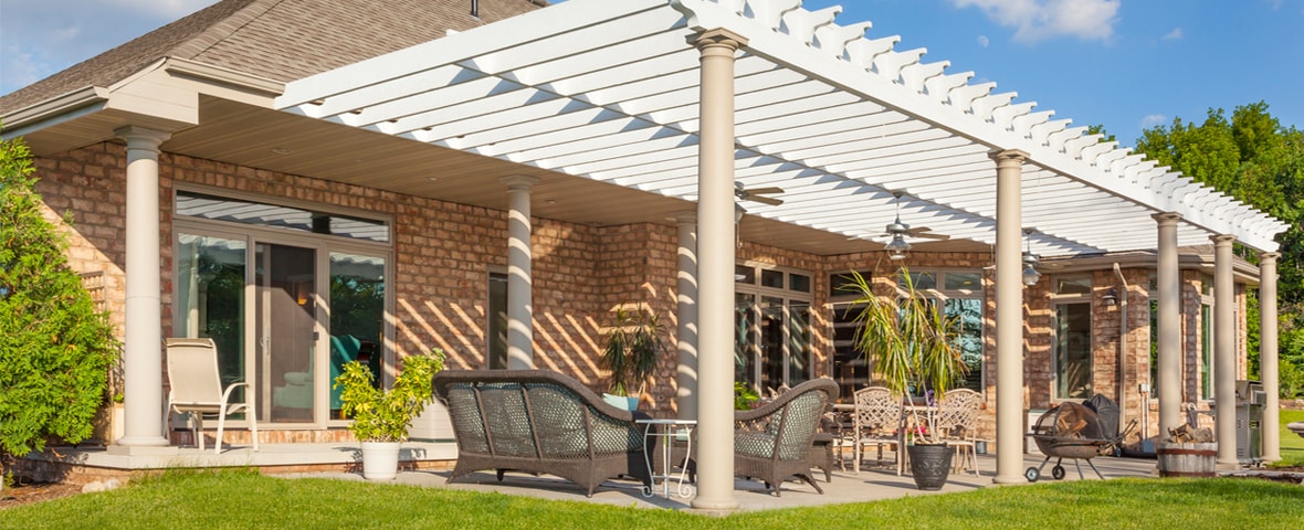Große überdachte Terrasse an einem Einfamilienhaus mit Ziegelfassade. Die Pergola mit weißen Lamellen spendet Schatten, darunter stehen Sitzgruppen, Pflanzenkübel und ein Grill.