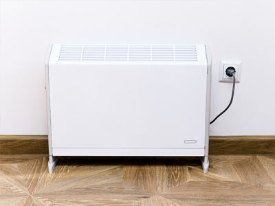Ein weißer elektrischer Heizkörper steht an einer Wand und ist über ein Kabel mit einer Steckdose verbunden. Der Boden besteht aus Holzparkett.