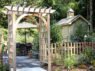 Ein Holzbogen mit Rankgitter markiert den Eingang zu einem gepflegten Gartenbereich mit Terrasse, Sitzgruppe, Sonnenschirm und Gartenhaus. Ein Holzlattenzaun und blühende Beete umrahmen die Szene.